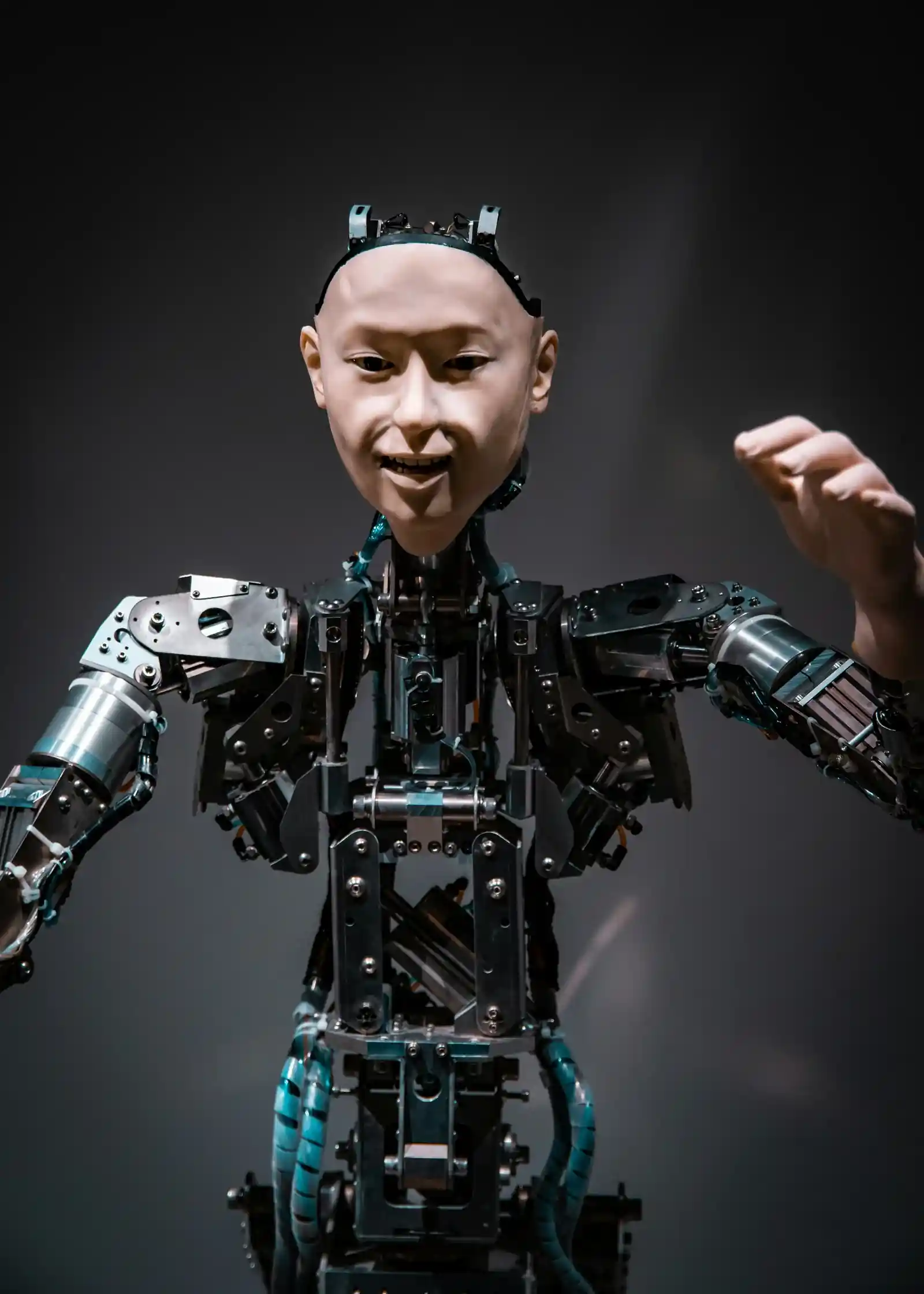 Robot con apariencia humana con razonamiento artificial, con el brazo izquierdo levantado a la altura de la cabeza, sobre fondo gris.